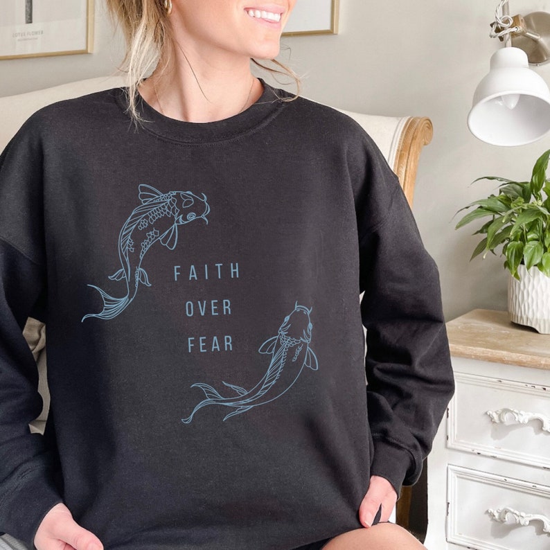 Puede incluir: Sudadera negra con un gr&aacute;fico de dos peces koi y el texto "FAITH OVER FEAR".