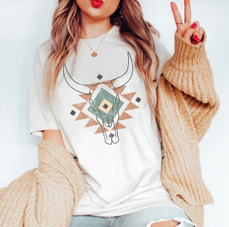 May include: A white t-shirt with a brown, green and white geometric design featuring a bull skull. The design is centred on the shirt.