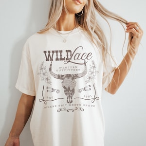 K&ouml;nnte beinhalten: Ein wei&szlig;es T-Shirt mit einem braunen Grafikdesign, das einen Stierkopf mit Spitzendetails, den Text "WILD Lace", "WESTERN OUTFITTERS", "EST. 1981" und "WHERE GRIT MEETS GRACE" zeigt.