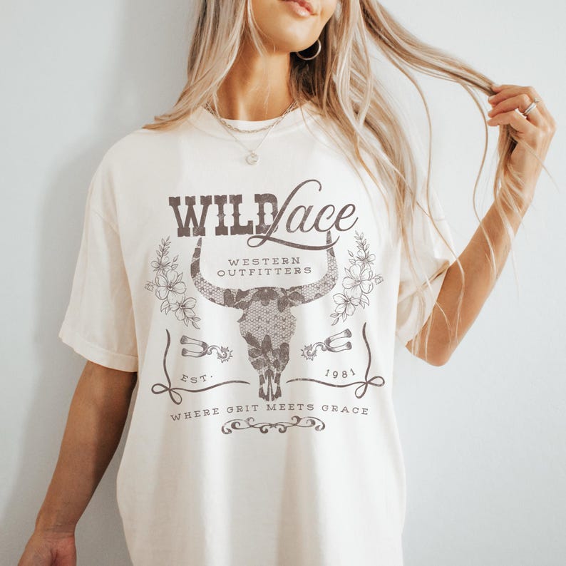 K&ouml;nnte beinhalten: Ein wei&szlig;es T-Shirt mit einem braunen Grafikdesign, das einen Stierkopf mit Spitzendetails, den Text "WILD Lace", "WESTERN OUTFITTERS", "EST. 1981" und "WHERE GRIT MEETS GRACE" zeigt.