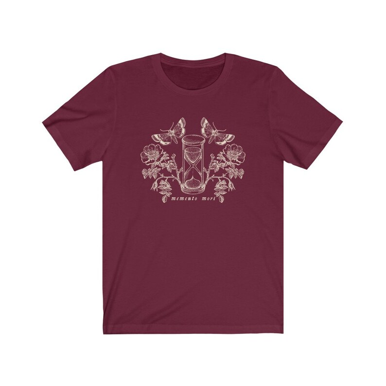 May include: A maroon t-shirt with a white graphic design of an hourglass, butterflies, and flowers. The text "Memento Mori" is printed below the hourglass.