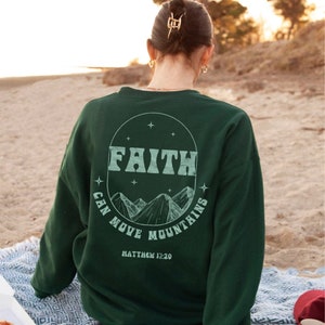 Puede incluir: Una sudadera verde oscuro con un gr&aacute;fico circular blanco en la espalda. El gr&aacute;fico presenta las palabras "FAITH CAN MOVE MOUNTAINS" y la escritura "MATTHEW 17:20".