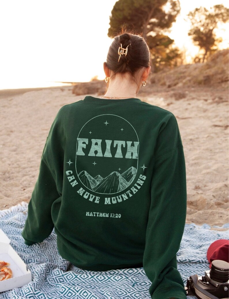 Puede incluir: Una sudadera verde oscuro con un gr&aacute;fico circular blanco en la espalda. El gr&aacute;fico presenta las palabras "FAITH CAN MOVE MOUNTAINS" y la escritura "MATTHEW 17:20".