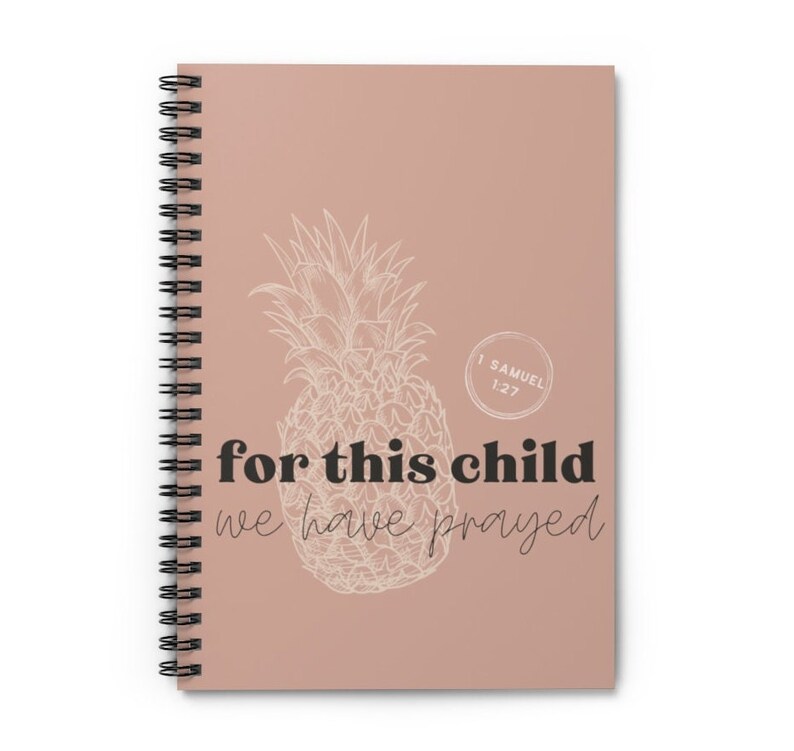 Puede incluir: Cuaderno de espiral con una cubierta marr&oacute;n claro. Presenta una ilustraci&oacute;n de pi&ntilde;a blanca y el texto "for this child we have prayed". Tambi&eacute;n se incluye el texto "1 Samuel 1:27". El cuaderno tiene una encuadernaci&oacute;n en espiral negra.