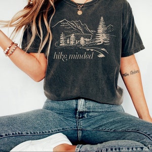 Op de afbeelding: Een donkergrijs T-shirt met een afbeelding van een bergketen en bomen. De tekst "hike minded" is onder de afbeelding gedrukt.
