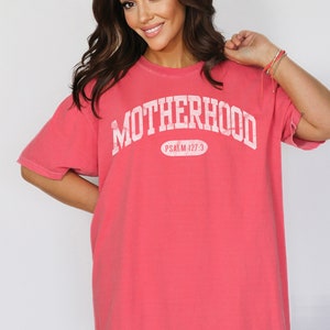 May include: A coral pink oversized t-shirt with the text "MOTHERHOOD" in white, arched across the front. The text "PSALM 127:3" is printed in white below the word "MOTHERHOOD".