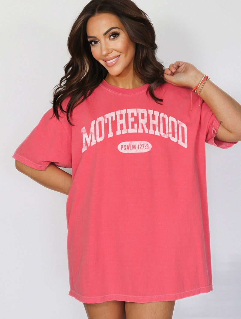 May include: A coral pink oversized t-shirt with the text "MOTHERHOOD" in white, arched across the front. The text "PSALM 127:3" is printed in white below the word "MOTHERHOOD".