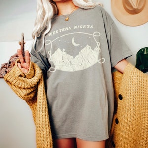 Może przedstawiać: Szara, oversize'owa koszulka z nadrukiem graficznym przedstawiającym pasmo g&oacute;rskie, sierp księżyca i sylwetkę kowboja. Tekst "WESTERN NIGHTS" jest nadrukowany w stylu vintage nad grafiką.