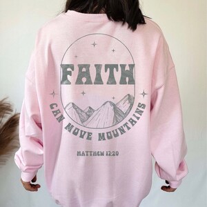 Puede incluir: Sudadera rosa con un estampado gr&aacute;fico de monta&ntilde;as y estrellas en un c&iacute;rculo. El texto "FAITH CAN MOVE MOUNTAINS" est&aacute; impreso en el c&iacute;rculo. El texto "MATTHEW 17:20" est&aacute; impreso debajo del c&iacute;rculo.