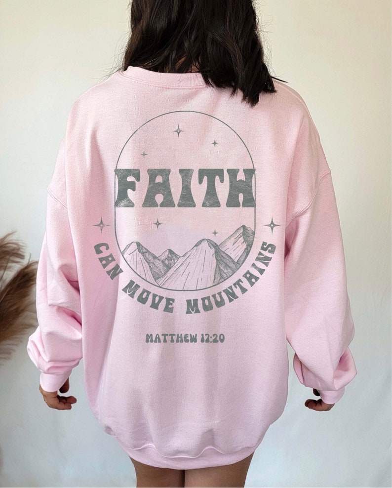 Puede incluir: Sudadera rosa con un estampado gr&aacute;fico de monta&ntilde;as y estrellas en un c&iacute;rculo. El texto "FAITH CAN MOVE MOUNTAINS" est&aacute; impreso en el c&iacute;rculo. El texto "MATTHEW 17:20" est&aacute; impreso debajo del c&iacute;rculo.
