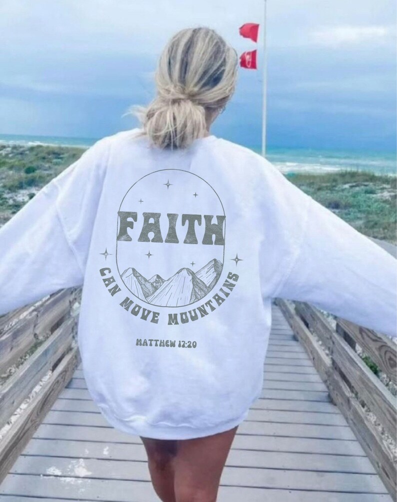 Puede incluir: Sudadera blanca con un estampado gr&aacute;fico de monta&ntilde;as y el texto "FAITH CAN MOVE MOUNTAINS" con la referencia b&iacute;blica "MATTHEW 12:20" debajo.