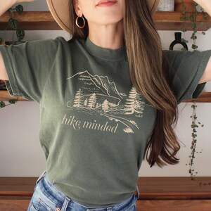 Op de afbeelding: Een groen T-shirt met een afbeelding van bergen en bomen. De tekst "hike minded" is op het shirt gedrukt.