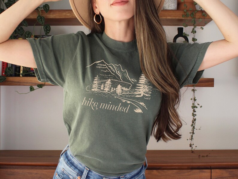 Op de afbeelding: Een groen T-shirt met een afbeelding van bergen en bomen. De tekst "hike minded" is op het shirt gedrukt.