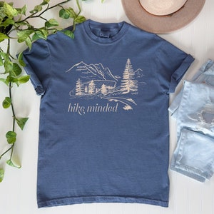 Op de afbeelding: Een blauw T-shirt met een witte afbeelding van een bergketen en bomen. De tekst "hike minded" is onder de afbeelding gedrukt.