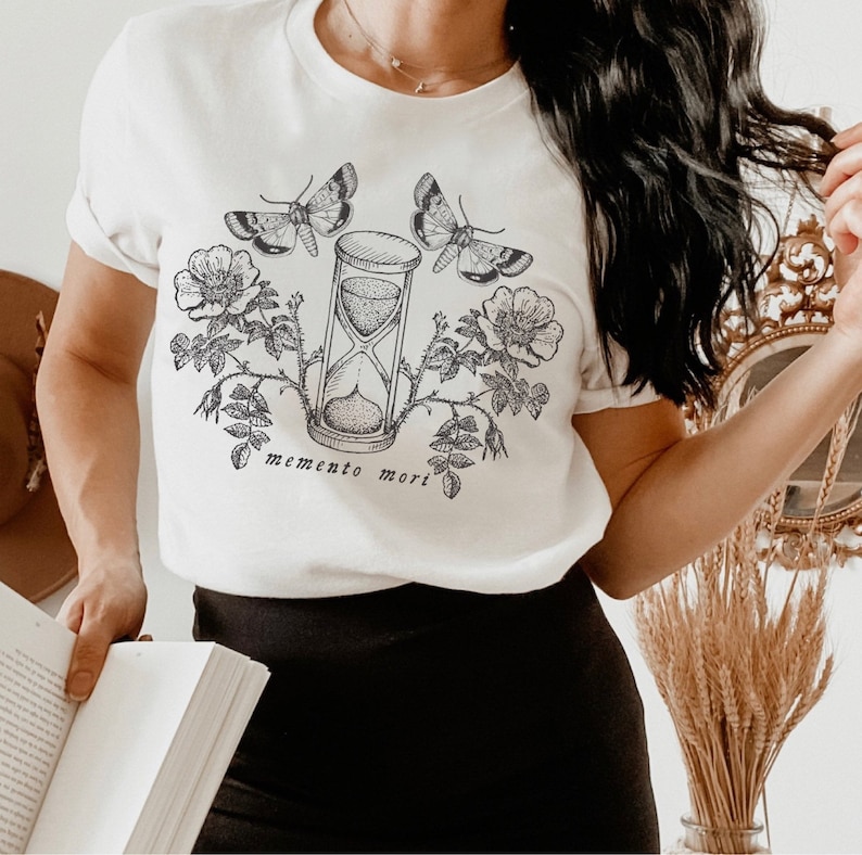 May include: White t-shirt with a black and white graphic design featuring an hourglass, butterflies, and roses. The text "memento mori" is printed below the hourglass.