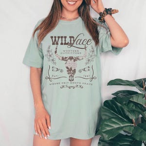 K&ouml;nnte beinhalten: Ein hellblaues T-Shirt mit einem braunen Grafikdesign, auf dem "Wild Lace Western Outfitters Est. 1981 Where Grit Meets Grace" steht. Die Grafik zeigt einen stilisierten Bullensch&auml;del mit floralen Akzenten.