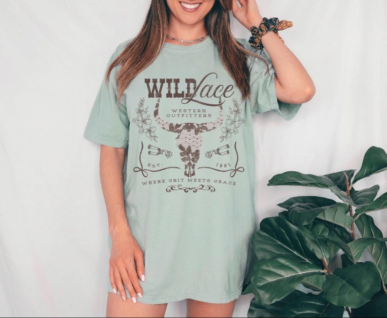 K&ouml;nnte beinhalten: Ein hellblaues T-Shirt mit einem braunen Grafikdesign, auf dem "Wild Lace Western Outfitters Est. 1981 Where Grit Meets Grace" steht. Die Grafik zeigt einen stilisierten Bullensch&auml;del mit floralen Akzenten.