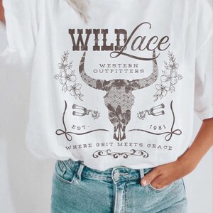 K&ouml;nnte beinhalten: Wei&szlig;es T-Shirt mit einem Grafikdesign eines Sch&auml;dels mit H&ouml;rnern und dem Text "Wild Lace Western Outfitters Est. 1981 Where Grit Meets Grace".