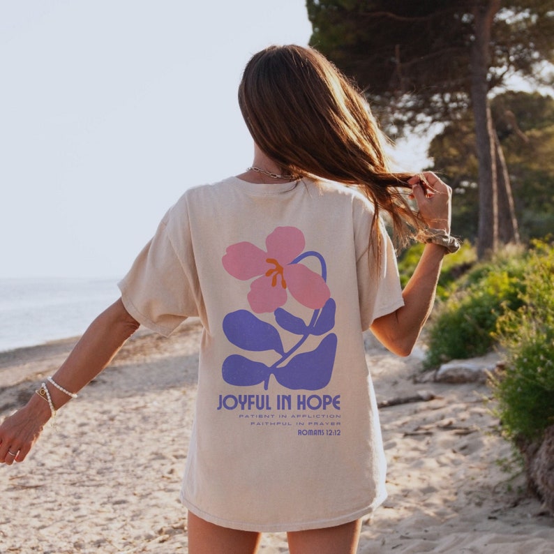 Op de afbeelding: Beige t-shirt met een bloemenontwerp op de achterkant. Het ontwerp heeft een roze bloem en blauwe bladeren, met de woorden "JOYFUL IN HOPE" en andere tekst eronder. Het shirt wordt gedragen door een persoon op een strand.