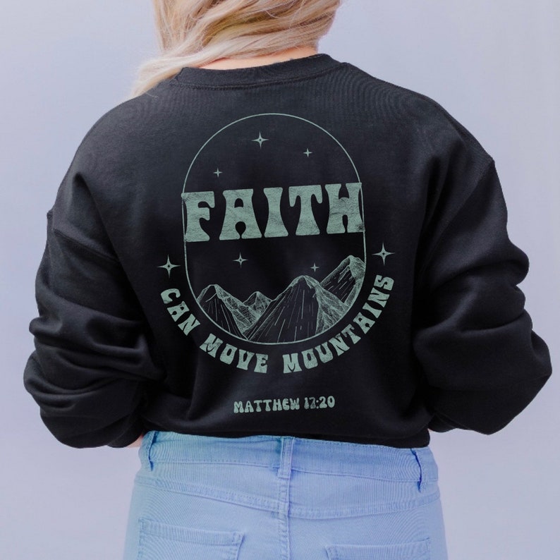 Puede incluir: Sudadera negra con un gr&aacute;fico circular verde en la espalda. El gr&aacute;fico presenta las palabras "FAITH CAN MOVE MOUNTAINS" y una cordillera con estrellas. El texto "MATTHEW 12:20" est&aacute; debajo del gr&aacute;fico.
