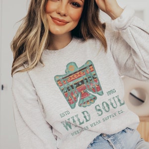 May include: A gray sweatshirt with a vintage-style graphic of a stylized eagle with geometric patterns in teal, red, orange, and black. The text "ESTD 1990 WILD SOUL WESTERN WEAR SUPPLY CO." is printed below the eagle.