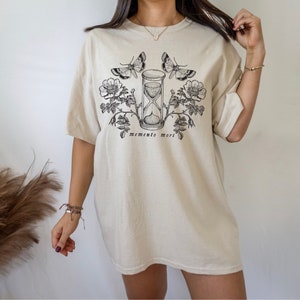 May include: A beige oversized t-shirt with a black graphic print of an hourglass, roses, and butterflies. The text "memento mori" is printed below the graphic.