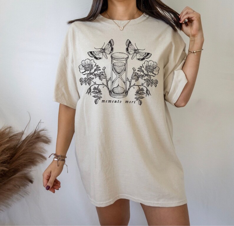 May include: A beige oversized t-shirt with a black graphic print of an hourglass, roses, and butterflies. The text "memento mori" is printed below the graphic.