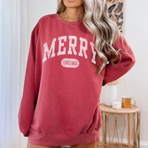 Op de afbeelding: Een rood sweatshirt met een witte afbeelding waarop "Merry Christmas" staat in een vintage universitaire lettertype.