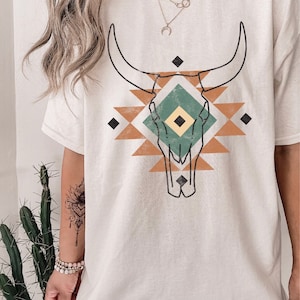 May include: A white t-shirt featuring a graphic print of a stylised bull skull within a geometric pattern, incorporating brown, green and yellow accents.
