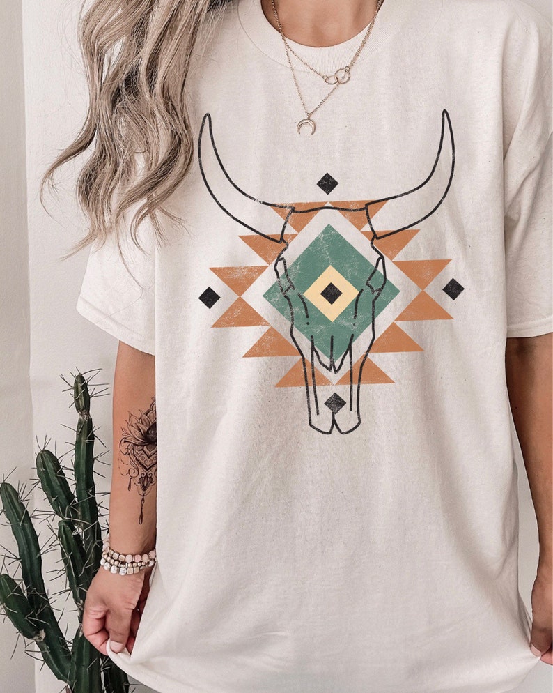 May include: A white t-shirt featuring a graphic print of a stylised bull skull within a geometric pattern, incorporating brown, green and yellow accents.