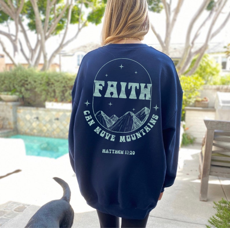Puede incluir: Sudadera azul marino con un gr&aacute;fico circular blanco en la espalda. El gr&aacute;fico presenta las palabras "FAITH CAN MOVE MOUNTAINS" y una cordillera con estrellas. El texto "MATTHEW 12:20" est&aacute; impreso debajo del gr&aacute;fico.