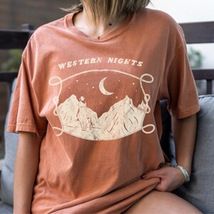 Może przedstawiać: Pomarańczowy t-shirt z nadrukiem graficznym przedstawiającym kowboja jadącego konno pod p&oacute;łksiężycem z gwiazdami. Tekst "WESTERN NIGHTS" jest nadrukowany nad grafiką.