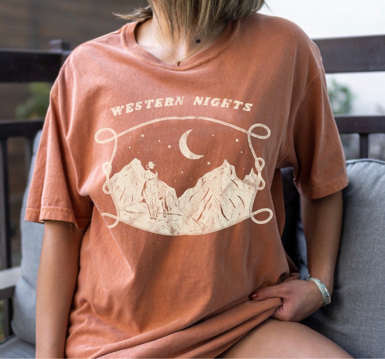 Może przedstawiać: Pomarańczowy t-shirt z nadrukiem graficznym przedstawiającym kowboja jadącego konno pod p&oacute;łksiężycem z gwiazdami. Tekst "WESTERN NIGHTS" jest nadrukowany nad grafiką.