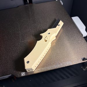 May include: A light beige 3D-printed tool, possibly a bottle opener or multi-tool, lies on a textured, dark surface. The tool has a distinctive shape with a curved handle, a notched edge, and several small holes. It features a decorative cross design.