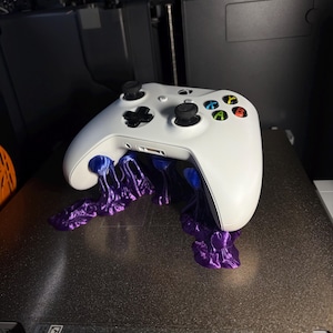 May include: A white Xbox controller with black joysticks and colored buttons, seemingly melting into a purple, tentacle-like structure. The controller is resting on a dark surface, possibly a 3D printer bed.