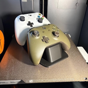 May include: Two Xbox controllers, one white and one olive green, rest on a black stand. The white controller is on top, with colorful buttons. The controllers are on a textured surface, with a 3D printer in the background.