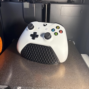 May include: A white Xbox controller with black accents and colored buttons. The controller features a black honeycomb pattern on the lower portion. The buttons are labeled with the letters X, Y, A, and B in blue, yellow, green, and red.