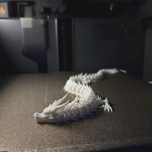 May include: A white, articulated dragon figurine, possibly 3D-printed, lies on a dark surface. The dragon features a detailed, spiky design and is in a curved pose. The background shows a 3D printer.