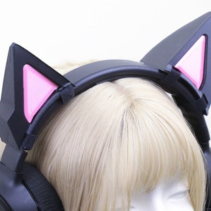 Puede incluir: Auriculares para juegos negros con orejas de gato. Las orejas de gato son negras con detalles rosas en el interior. Los auriculares están sobre una peluca rubia. Las orejas de gato están sujetas a los auriculares con correas de velcro negras.
