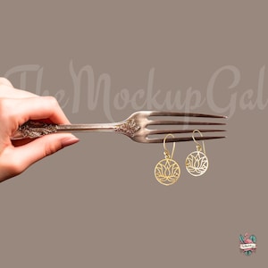 May include: A pair of gold-toned earrings with a delicate lotus flower design. The earrings are hanging from a silver fork.
