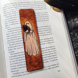 May include: A wooden bookmark with a ghost illustration holding flowers. The ghost is white with black eyes and a black outline. The bookmark has a textured, brown background and is resting on an open book.