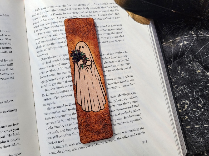 May include: A wooden bookmark with a ghost illustration holding flowers. The ghost is white with black eyes and a black outline. The bookmark has a textured, brown background and is resting on an open book.