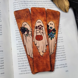 May include: Three Halloween-themed bookmarks with a distressed leather texture. Each bookmark features a white ghost with floral accents and a pumpkin. The bookmarks are resting on an open book.