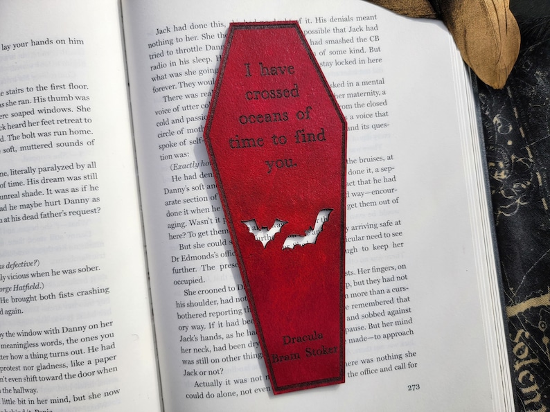 May include: A red wooden coffin-shaped bookmark with a quote from Dracula by Bram Stoker. The quote reads "I have crossed oceans of time to find you." Two bats are cut out of the bookmark.