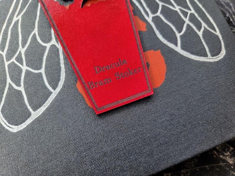 May include: A red book cover with a white insect design and a red coffin-shaped bookmark with the text "Dracula Bram Stoker" in black.