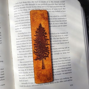 May include: A brown leather bookmark with a detailed illustration of a pine tree. The bookmark is worn and has a rustic look. The text "Exactly how dangerous it was" is visible on the page behind the bookmark.