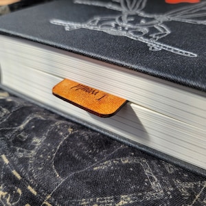 May include: A black hardcover book with a silver embossed design on the cover. A brown leather bookmark with the text "From 1" is attached to the book.