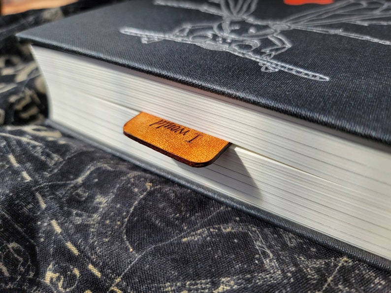 May include: A black hardcover book with a silver embossed design on the cover. A brown leather bookmark with the text "From 1" is attached to the book.