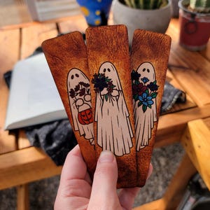 May include: Three wooden bookmarks with a Halloween ghost theme. Each bookmark features a white ghost with different floral arrangements and a pumpkin. The bookmarks are held in a hand.