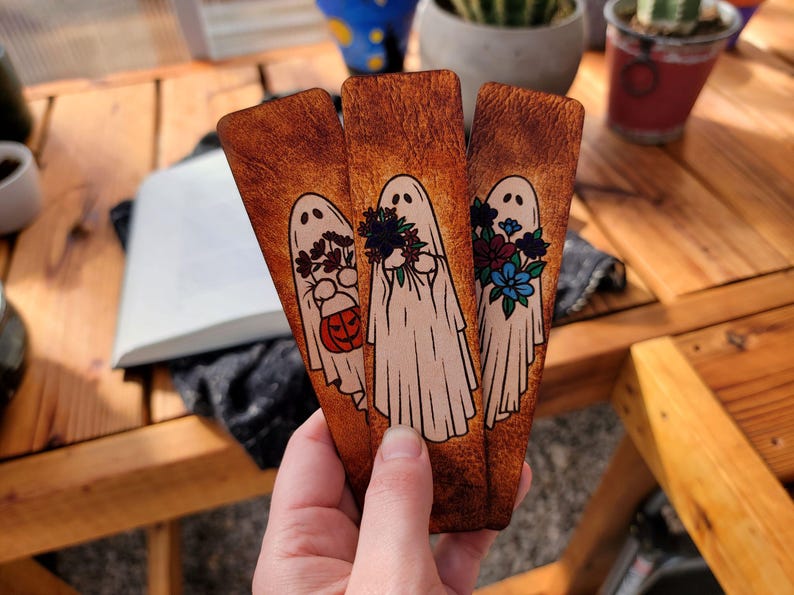 May include: Three wooden bookmarks with a Halloween ghost theme. Each bookmark features a white ghost with different floral arrangements and a pumpkin. The bookmarks are held in a hand.